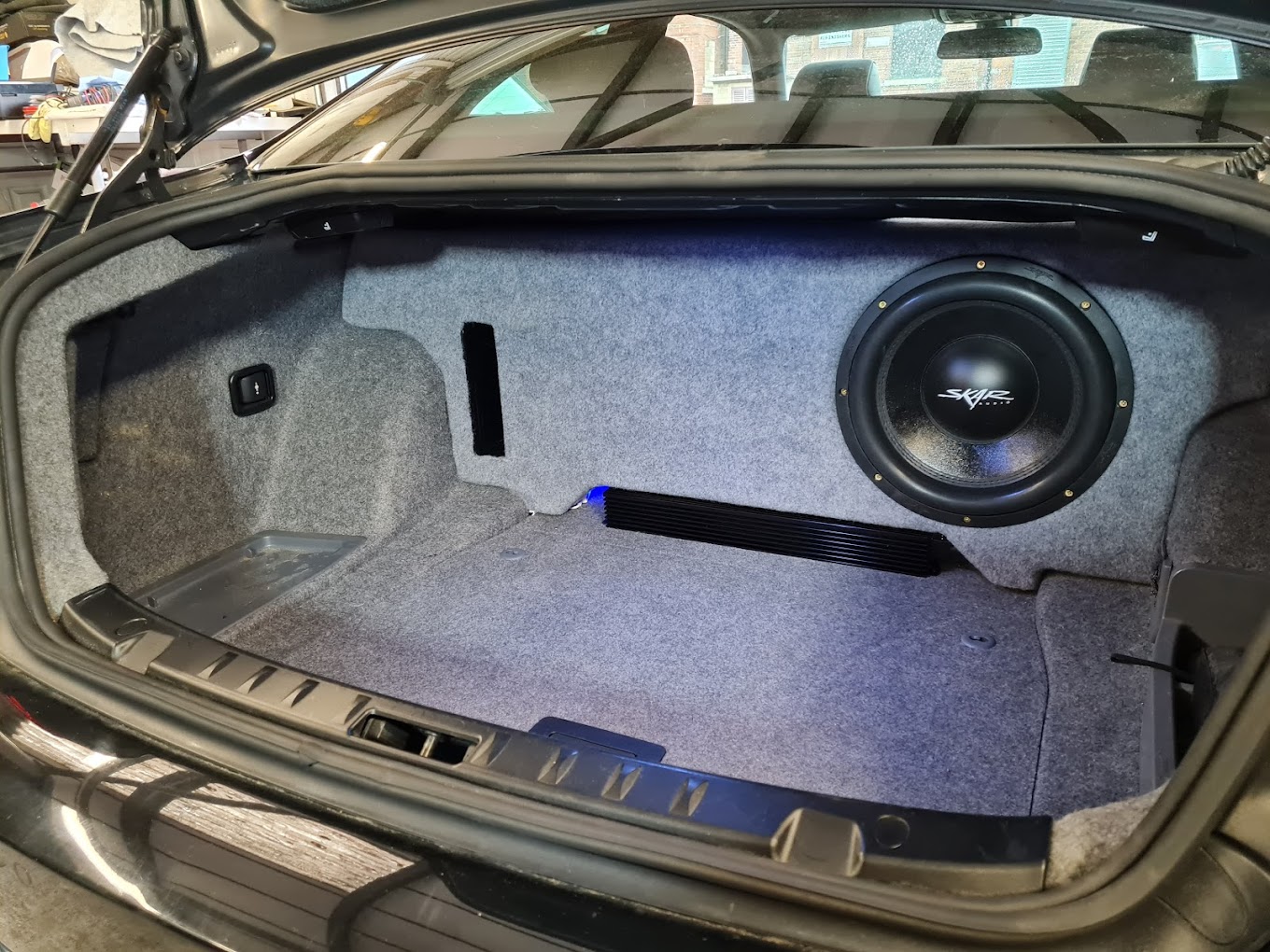 A single SKAR subwoofer set into a carpeted enclosure in a car boot, with the amplifier racked below and the trim refitted
